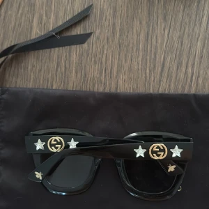 Svarta Gucci GG0208S solglasögon med stjärnor - Säljer ett par svarta Gucci GG0208S solglasögon med bred båge, silvriga stjärnor och små guldfärgade bin på sidan. Snygg, markerad form som ger en lyxig vibe. Kommer med originalpåse från Gucci.