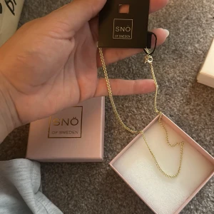 Guldfärgat halsband från SNÖ of Sweden - Stilrent guldfärgat halsband från SNÖ of Sweden med tunn kedja och klassisk karbinlås. Kommer i original ask, perfekt för dig som gillar minimalistiska smycken med en touch av lyx. Det är helt oanvänt 