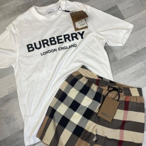 Vit Burberry t-shirt med logga shorts ingår  - Vit t-shirt från Burberry med stor svart logga och texten 'London England' på bröstet. Klassisk rund hals och korta ärmar. Tillverkad i mjuk bomull som känns skön mot huden. Perfekt för dig som gillar stilrena och exklusiva plagg. Shorts ingår 