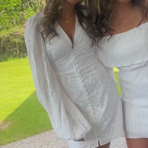 populär vit ADOORE klänning - Superfin vit kort klänning med v-ringning, puffiga långärmar och knappar framtill. Klänningen har broderade blommor i tyget och markerad midja för en snygg siluett. Perfekt för student eller sommarens alla tillfällen!