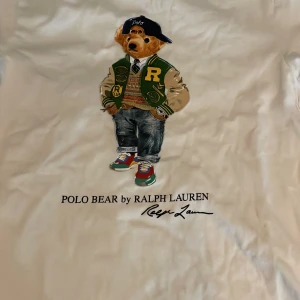 Vit Polo Bear t-shirt från Ralph Lauren - Vit t-shirt från Ralph Lauren med ikoniskt Polo Bear-tryck framtill. Klassisk passform med rund hals och korta ärmar. Trycket visar en nalle i streetwear-stil och texten 'POLO BEAR by RALPH LAUREN'. Skön bomullskvalitet, perfekt för en avslappnad look. Använt ett ental gånger 