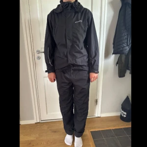 Svart vind/regn-jacka och byxor från Everest 158/164 - Svart vind/regn-jacka och byxor från Everest i storlek 158/164. Jackan har huva, dragkedja framtill och justerbara ärmslut. Materialet är lätt och vattenavvisande polyester, perfekt för blåsiga dagar. Snygg och enkel design med diskret logga på bröstet. Byxorna har reså i midja och vid anklarna.