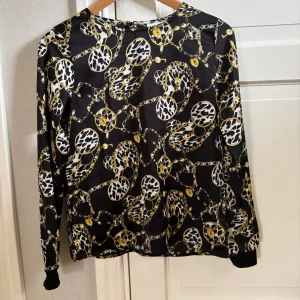 Svart blus med kedje- och leopardmönster - Snygg svart blus från Grunt i storlek L med unikt kedje- och leopardmönster i vitt och gult. Blusen har långa ärmar med muddar och är tillverkad i ett glansigt, mjukt material som känns lyxigt. Perfekt för dig som vill sticka ut med coola detaljer. Barnstorlek så skulle säga 158/164.