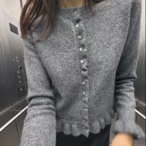 Grå kofta med volangkant och knappar - Superfin grå kofta i mjukt stickat material med volangkant nertill och längs ärmsluten. Stängs med små knappar framtill och har en klassisk rund halsringning. Perfekt att slänga över en topp för en chill look.