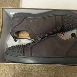 Mörkgrå Christian Louboutin sneakers - Snygga mörkgrå sneakers från Christian Louboutin i mocka med ikoniska röda sulor och coola nitar på tån. Skorna har snörning och en platt sula, perfekt för dig som vill sticka ut med en edgy och lyxig stil.