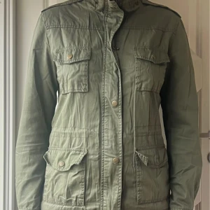 Grön parkajacka från Bik Bok - Militärgrön vår/sommarjacka från Bik Bok i 100% bomull.. Strl S. Jackan har fyra stora fickor framtill, axelklaffar och stängs med både dragkedja och knappar.  Dragsko i midjan på insidan av jackan (se bild) . Använd men i bra skick.
