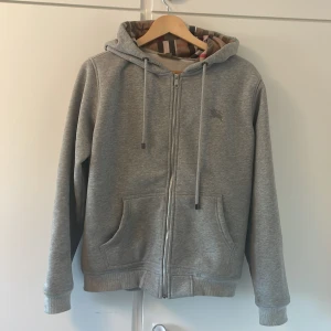 Grå hoodie från Burberry med dragkedja - Snygg grå hoodie från Burberry i bomull med klassiskt rutigt foder i huvan. Tröjan har dragkedja, snörning i huvan, broderad logga på bröstet och två stora fickor framtill. Perfekt för dig som gillar stilrena och exklusiva plagg.