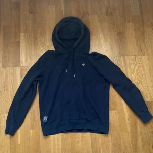 Mörkblå hoodie från Morris - Säljer en mörkblå hoodie från Morris med klassisk huva och dragsko. Tröjan har en liten broderad logga på bröstet och ribbade muddar vid ärmslut och nederkant. Perfekt för en chill och stilren look.