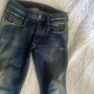 Blå Replay jeans med slitningar - Snygga blå jeans från Replay med coola slitningar och tvättade detaljer. Klassisk femficksmodell med kontrastsömmar och läderpatch bak. Jeansen har slim passform och är tillverkade i bomull. Perfekta för en avslappnad look. En liten defekt (sida2) därav priset. 