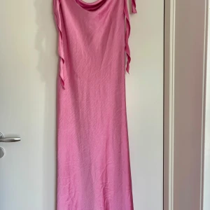 Rosa långklänning med volangdetaljer - Säljer en snygg rosa långklänning med volangdetaljer längs axelbanden och sidorna. Klänningen har en mjuk draperad urringning och är ärmlös, vilket ger en chill och feminin vibe. Perfekt för sommarens alla tillfällen när du vill sticka ut lite extra. Rosa silkesklänning, lång. Storlek 40, från ginatricot. Använd en gång!
