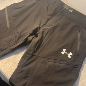 Svarta Under Armour byxor - Skriv dm för video/prisförslag! Säljer ett par svarta täckbyxor från Under Armour med stilren design och logga på benet. Byxorna har flera praktiska dragkedjefickor och knappstängning i midjan. Materialet är slitstarkt syntet, perfekt för utomhusaktiviteter och sport.