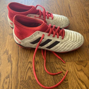 Adidas Predator fotbollsskor röd/vit - Adidas Predator fotbollsskor i rött och vitt med svarta detaljer. Skorna har snörning, ribbad textil vid ankeln och en platt sula med dobbar för gräsplan. Snygg design med klassiska tre svarta ränder på sidan och Predator-tryck vid hälen. Perfekt för dig som vill sticka ut på planen. Ingen låda tyvärr 