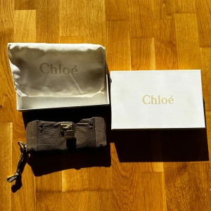 Brun plånbok från Chloé i skinn - Snygg brun plånbok från Chloé i äkta skinn med coolt lås och nyckeldetalj i metall. Plånboken har dragkedja och flera fack för kort och kontanter. Kommer med original dustbag och box. Perfekt för dig som gillar lyxiga accessoarer.