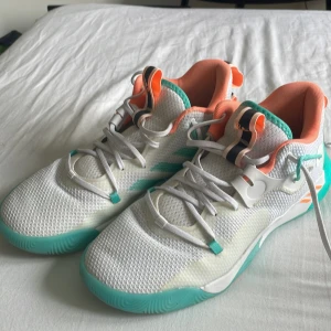  James Harden basket skor - Fräscha Adidas sneakers med mesh-ovandel i vitt, turkos sula och detaljer i orange och beige. Skorna har snörning och rund tå, samt coola mönster på yttersulan. Perfekt för dig som gillar sportig stil och färgglada detaljer.