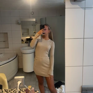 Beige ribbad kortklänning med knyt - Superfin beige ribbad klänning med lång ärm och knyt i midjan. Snygg, figurnära passform och mjukt stickat material som sitter skönt.  Använd en gång 