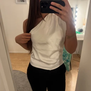 Vit oneshoulder topp i satin - Säljer en stilren vit oneshoulder topp i satinliknande material. Toppen har en snygg asymmetrisk skärning och är ärmlös på ena sidan, vilket ger en modern och trendig look. Perfekt för dig som vill sticka ut med en enkel men elegant stil.