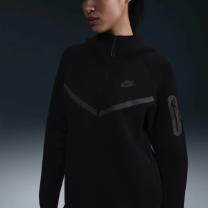 Svart Nike tech - Nike hoodiejacka i svart med hel dragkedja, huva och diskret logga på bröstet. Snygg V-formad detalj framtill och praktisk ficka med dragkedja på ärmen. Perfekt för en sportig och clean look.
