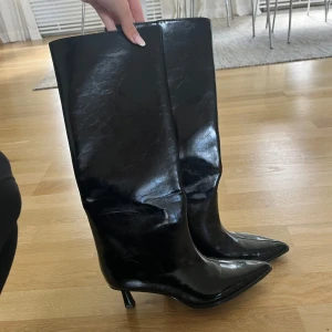 Svarta stövlar från Zara - Säljer ett par svarta, glansiga stövlar från Zara med spetsig tå och smal klack. Stövlarna är höga och har en stilren, minimalistisk design som ger en edgy vibe. Materialet är syntet med skinnlook och de passar perfekt till att lyfta din outfit. Endast använda en gång