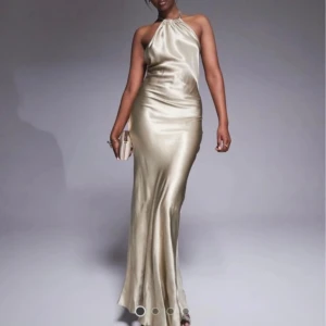 Champagnefärgad lång halterneckklänning - Säljer en elegant långklänning i champagnefärgad satin med halterneck och slimmad passform. Klänningen har en mjukt skimrande yta och faller snyggt längs kroppen. Perfekt för bal eller student. Materialet är glansigt och känns lyxigt mot huden. Använd en gång💕
