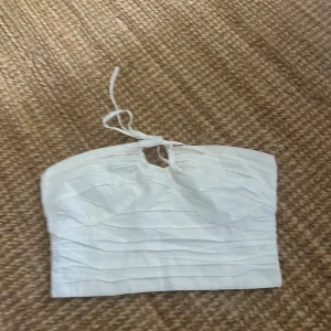 Vit bandeau tubtopp med knyt - Säljer en vit bandeau tubtopp med snygg knytning och liten öppning framtill. Toppen har en lätt rynkad struktur på framsidan och smockad baksida. Toppen är ganska kort