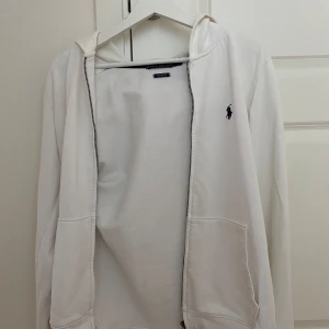 Vit hoodie från Polo Ralph Lauren - Vit hoodie från Polo Ralph Lauren med klassisk blå broderad logga på bröstet. Tröjan har dragkedja, huva och två fickor framtill. Materialet är mjuk bomull och passformen är custom fit. Perfekt för chill dagar och enkel att matcha med jeans eller joggers.