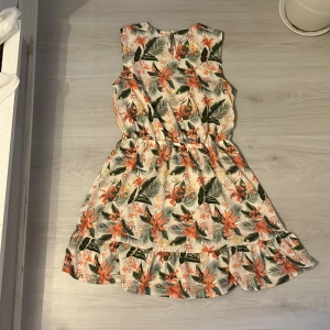 Blommig kortklänning från Basic Needs - Superfin kortklänning med tropiskt blommigt mönster i vitt, orange, grönt och lite gult. Klänningen är ärmlös, har volangkant nertill och ett knytband i midjan för extra snygg passform. Perfekt för sommarens alla häng och utflykter!
