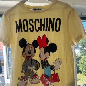 HM x MOSCHINO T-shirt - Säljer en gul t-shirt från Moschino i samarbete med HM. Framsidan har stort Moschino-tryck och motiv med Mickey och Minnie Mouse i streetwear. T-shirten har rund hals och korta ärmar, perfekt för dig som gillar statement-plagg. Aldrig använd.