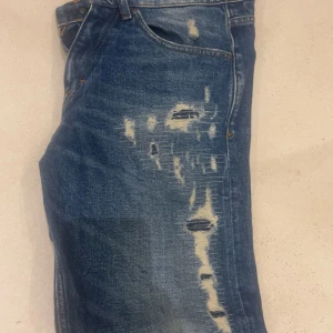 Tiger of Sweden blå slitna jeans - Säljer ett par blå jeans från Tiger of Sweden med snygga slitningar och hål på framsidan. Klassisk femficksmodell med raka ben och normal passform. Jeansen har en mörkblå tvätt och är tillverkade i bomull. Perfekt för dig som gillar en avslappnad och sköna jeans.Pris kan diskuteras vid snabb affär. Hör av er!