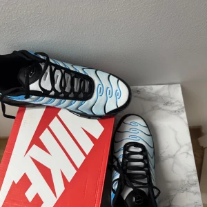 Nike Air Max Plus blå/svart EUR 42 - Nike Air Max Plus sneakers i en fräsch ljusblå och svart färgkombination med vågiga mönster och svarta detaljer. Skorna har synliga Air Max-enheter i sulan, snörning och en mesh-överdel som ger en sportig vibe. Perfekt för dig som gillar unika och stilrena sneakers.
