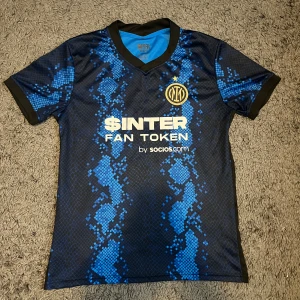 Inter Milan blå fotbollströja - Snygg Inter Milan fotbollströja med svart och blått ormmönster, guldigt klubbmärke och vit sponsorlogga på bröstet. Tröjan har korta ärmar, V-ringad krage med svart kant och är gjord i lätt, ventilerande polyester. Perfekt för match eller träning!