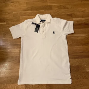 Vit Polo Ralph Lauren t-shirt - Vit klassisk t-shirt från Polo Ralph Lauren med korta ärmar och krage. T-shirten har tre knappar framtill och den ikoniska broderade loggan i marinblått på bröstet. Materialet är mjuk bomull och passformen är normal. Perfekt för en clean och stilren look.