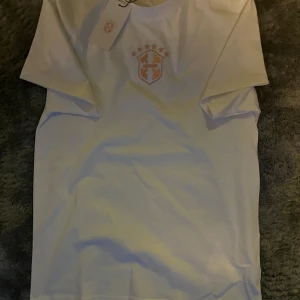 Vit t-shirt med broderad logga - Fräsch vit t-shirt från Avenir med snygg broderad logga i rosa och vitt på bröstet. Klassisk passform med rund hals och korta ärmar. Perfekt för dig som gillar stilrena plagg med en twist. Materialet känns mjukt och skönt mot huden.