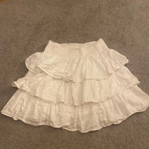 Vit volangkjol med resårmidja - Supersöt vit kortkjol med flera lager volanger och bred smockad resår i midjan. Kjolen har en lätt skrynklig struktur och ger en riktigt flowy känsla. storlekn är 146-152 men eftersom att den är stretchig så skulle jag säga XS. Skriv privat för bilder på💕lite skrynklig men stryker innan leverans!