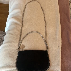 Svart axelväska med kedja och dragkedja - Snygg svart axelväska i mocka med silverfärgad kedja och dragkedja. Väskan har en rund metalltag och detaljerad kant med flätad mocka. Perfekt storlek för det viktigaste och cool design som sticker ut. Den är helt oanvänd, så i topp skick! Kan självklart skicka fler bilder, vet tyvärr inte vart den är ifrån