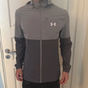 Grå vindjacka från Under Armour - Snygg tvåfärgad vindjacka från Under Armour i ljusgrått och mörkgrått. Jackan har huva, hel dragkedja framtill och en diskret logga på bröstet. Perfekt för dig som gillar sportig stil och vill ha något lätt och funktionellt.