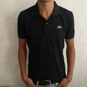 Svart Lacoste pikétröja i bomull - Svart pikétröja från Lacoste med klassisk krage och korta ärmar. Tröjan har den ikoniska gröna krokodilloggan broderad på bröstet och är gjord i mjuk bomull. Passformen är normal och stilen är clean och tidlös.