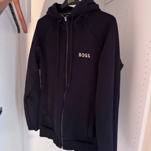 Svart hoodie från BOSS med dragkedja - Svart hoodie från BOSS med guldfärgad logga på bröstet. Tröjan har huva med snörning, hel dragkedja framtill och långa ärmar. Tillverkad i mjuk bomull, perfekt för chill dagar. Snygg och enkel design som funkar till det mesta.