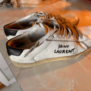 Saint Laurent skor äkta - Säljer ett par vita sneakers från Saint Laurent med handskriven logga på sidan. Skorna har svarta detaljer vid hälen, rund tå och platt sula. Tillverkade i skinn med beige gummisula och klassisk snörning. Perfekt för dig som gillar streetstyle och designer.