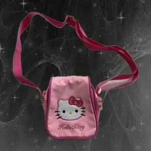 Hello Kitty rosa mini axelväska - Söt rosa axelväska med broderat Hello Kitty-ansikte och text på framsidan. Väskan har justerbar axelrem, magnetknäppning och ett fack med dragkedja på baksidan. Tillverkad i slitstark polyester med detaljer i olika rosa nyanser och en liten Hello Kitty-etikett vid sidan. En liten fläck- se bild 2. 