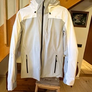 Ljusgrå vindjacka från Wedze S - Snygg ljusgrå och vit vindjacka från Wedze med huva och svarta dragkedjor. Jackan har tre fickor med dragkedja och justerbara ärmslut med kardborre. Perfekt för dig som gillar stilren och sportig look. Vattentät och andas, designad för alpina äventyr.