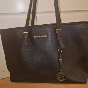 Svart axelväska från Michael Kors - Stilren svart axelväska från Michael Kors med guldfärgade detaljer och MK-hänge. Väskan har flera fack och dragkedja på insidan, perfekt för att hålla ordning på dina grejer. Tillverkad i skinn med strukturerad yta och långa axelremmar. INTE SÄKER om den är äkta för jag köpte den på sellpy men den kan va🥰