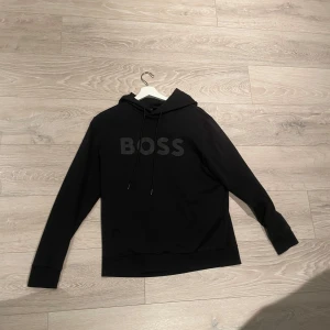 Svart hoodie från BOSS med reflektiv logga - Svart hoodie från BOSS med stor vit logga på bröstet. Tröjan har huva med snörning, lång ärm och en klassisk känguruficka framtill. Mjuk och skön bomullskvalitet som passar perfekt för chill eller streetwear. Snygg och enkel design med loose passform. PRIS KAN DISKUTERAS!