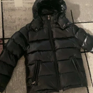 Svart Moncler dunjacka med huva - Säljer en svart, glansig dunjacka från Moncler med huva och två dragkedjefickor framtill. Jackan har Moncler-logga på ärmen och quiltad design. Perfekt för dig som vill ha en varm och snygg jacka med streetkänsla. Dragkedja framtill och cool pufferstil.