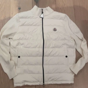 Vit moncler cardigan - Snygg vit pufferjacka från Moncler med ribbad krage och ärmar. Jackan har svart dragkedja, två fickor med knappar och Moncler-logga på bröstet. Materialet är en mix av polyester och stickad ull, vilket ger en skön och modern look.