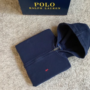 Marinblå hoodie från Polo Ralph Lauren - Snygg marinblå hoodie från Polo Ralph Lauren med dragkedja och huva. Tröjan har det klassiska röda Polo-logot broderat på bröstet och är tillverkad i mjuk bomull. Perfekt för chill dagar och enkel att matcha med jeans eller joggers.