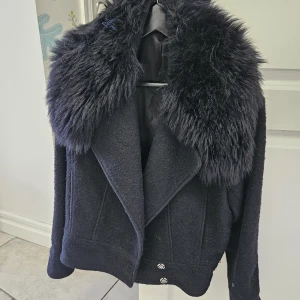 Svart pälsjacka från Filippa K - Snygg svart pälsjacka från Filippa K med stor fluffig krage i fuskpäls och coola detaljer som dubbla spännen vid midjan. Jackan har breda slag, knappar framtill och är helfodrad med mjukt tyg. Perfekt för dig som vill ha en statementjacka med lyxig vibe.