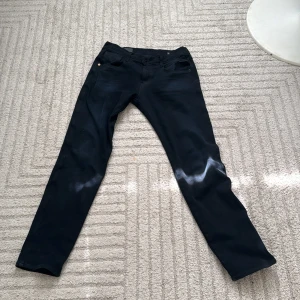 Replay Hyperflex svarta jeans, stl 30 - Svarta Replay Hyperflex jeans med klassisk femficksdesign och snygga detaljer vid fickorna. Stretchigt material som sitter skönt och följer kroppen. Passformen är slim och byxorna har en rak siluett. Perfekta för dig som gillar stilrena och moderna jeans.