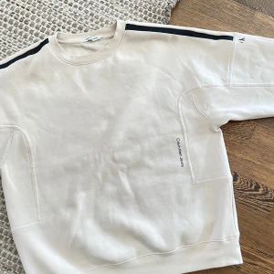 Beige sweatshirt från Calvin Klein - Snygg beige sweatshirt från Calvin Klein Jeans med svart logga på ärmen och diskret tryck på sidan. Tröjan har svarta ränder längs axlarna och en rund hals. Perfekt för en chill och stilren look, passar till vadsomhelst
