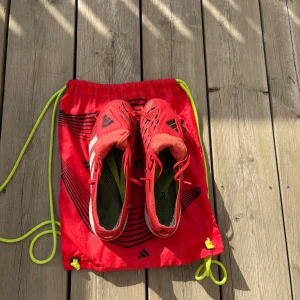 Adidas Predator röda fotbollsskor - Säljer ett par Adidas Predator fotbollsskor i röd och vit färg med svarta detaljer. Skorna har snörning, platt sula med dobbar och klassiska Adidas-ränder på sidan. Ovandelen är i syntetmaterial och designen är riktigt sportig och modern. Perfekt för dig som vill sticka ut på planen.