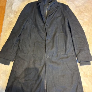 Svart rock från Smog, storlek L - Snygg svart rock från Smog i storlek L. Klassisk design med krage, knappar framtill och två stora fickor. Rocken har ribbade muddar vid ärmslut och är tillverkad i ett mjukt material som känns lyxigt. Perfekt för dig som gillar stilrena och tidlösa plagg.